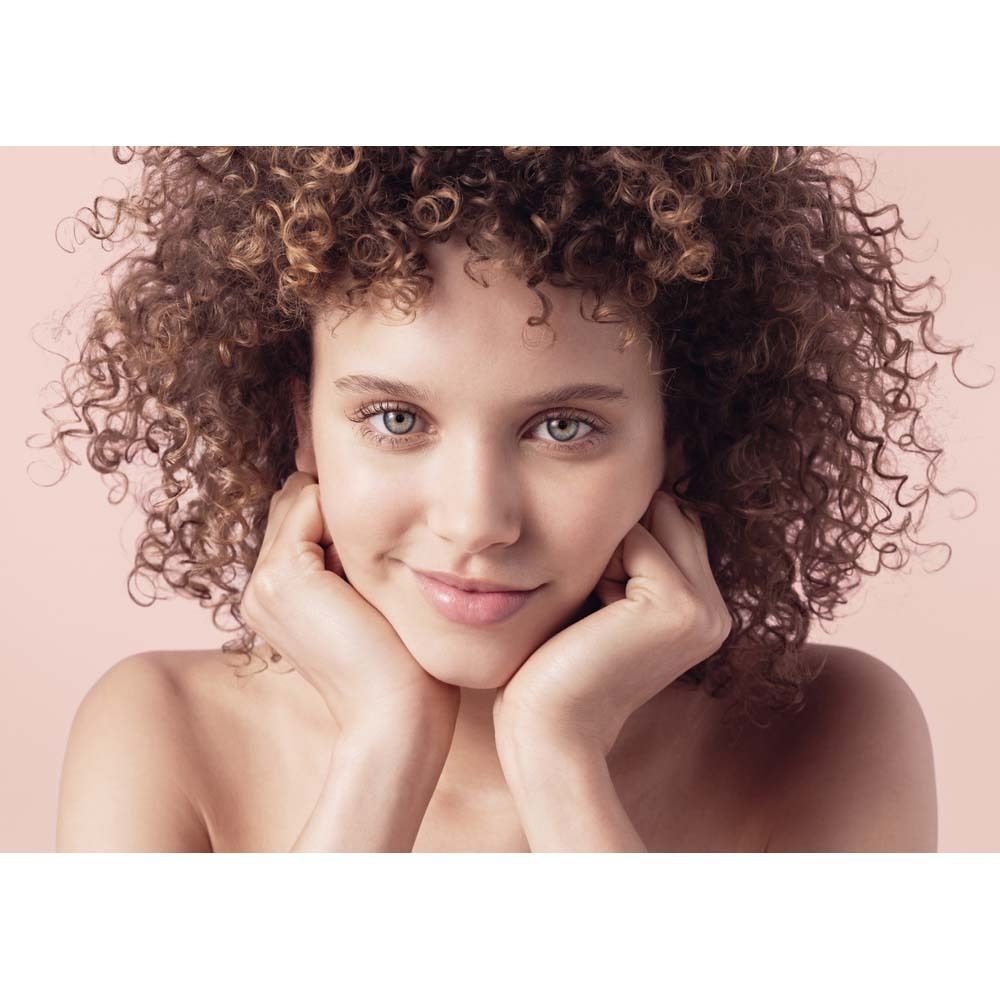 Portrait d'une femme aux cheveux bouclés. Elle tient ses mains sur son visage. Fond rose.