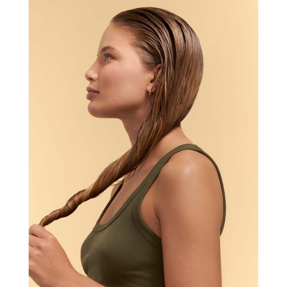 Femme avec cheveux mouillés tressés. Elle porte un débardeur vert olive. Fond jaune.