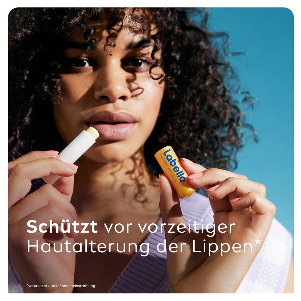 Jeune femme aux cheveux bouclés foncés appliquant du baume à lèvres Labello SUN PROTECT, tenant le stick.