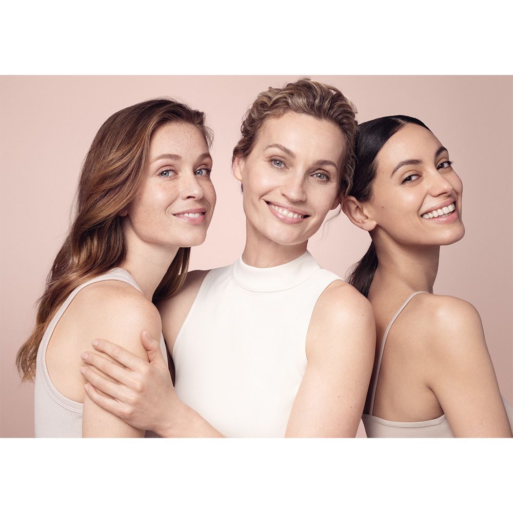 Trois femmes souriantes sur fond rose. Elles portent des hauts clairs. Différentes couleurs de cheveux et de peau.
