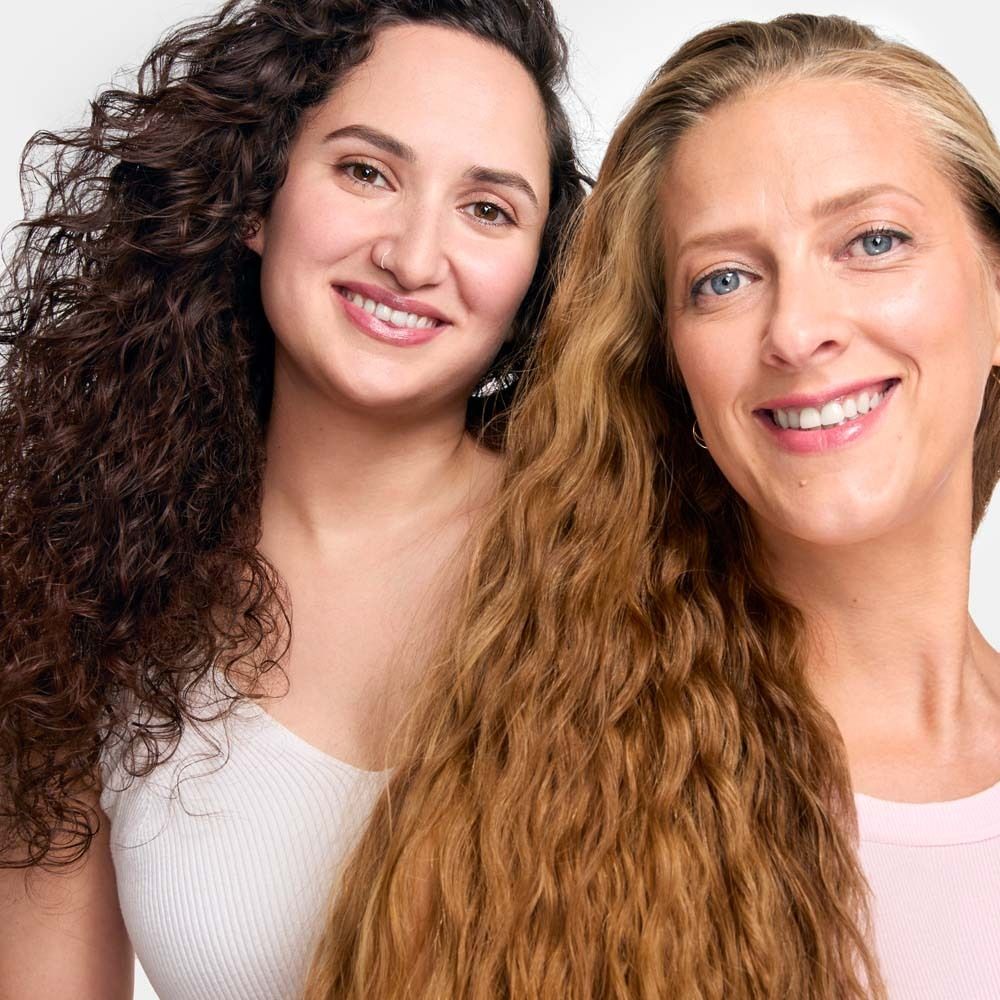 Deux femmes avec des cheveux bouclés et ondulés. Elles sourient à la caméra. Une femme a les cheveux bruns, l'autre blonds.