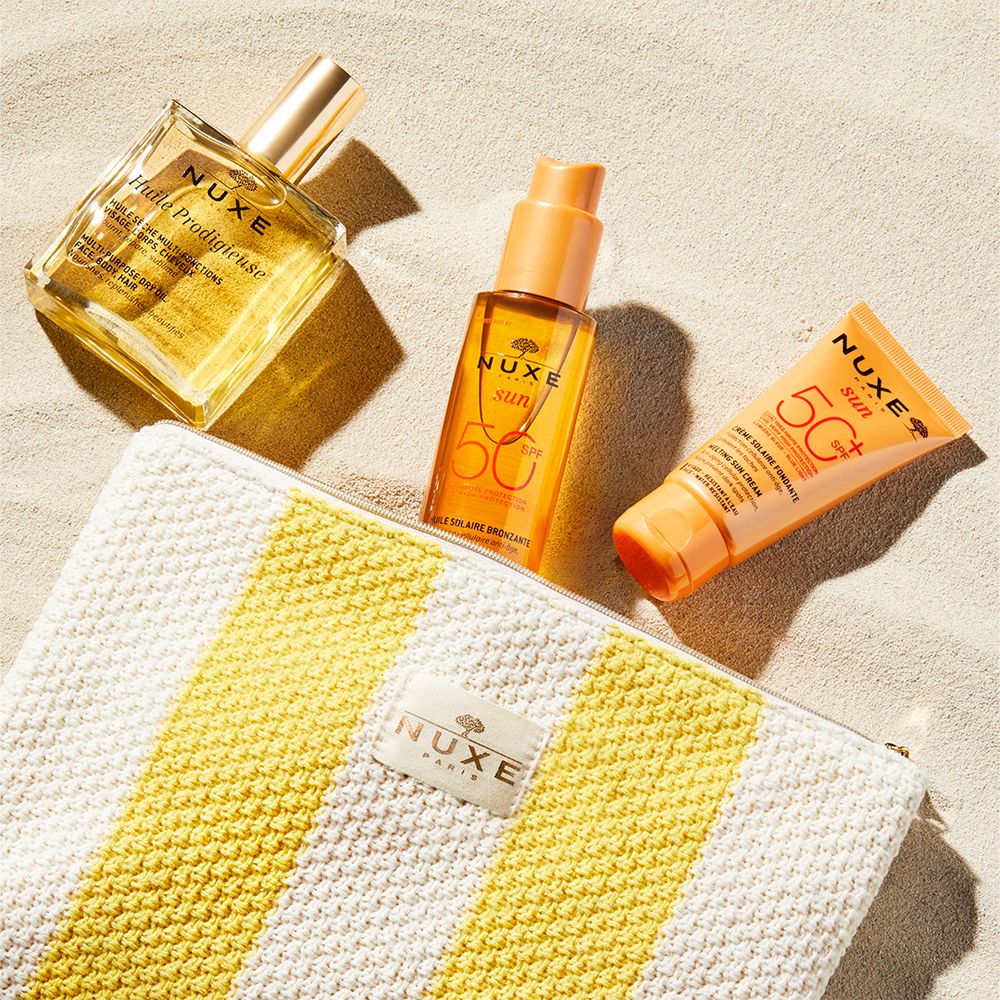 Huile solaire, crème solaire et flacon d'huile sur du sable. Une trousse rayée jaune est à côté.