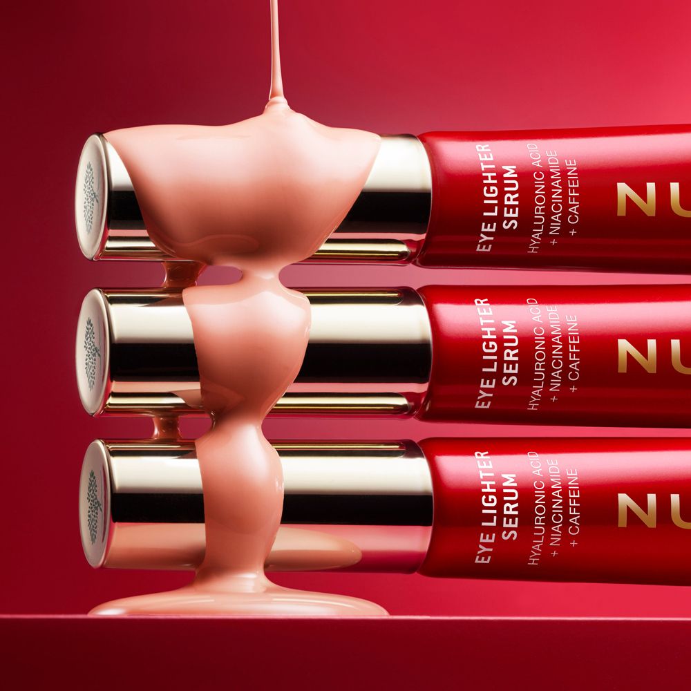 Drei Tuben NUXE Eye Lighter Serum. Rosa Flüssigkeit wird auf die Tuben gegossen.