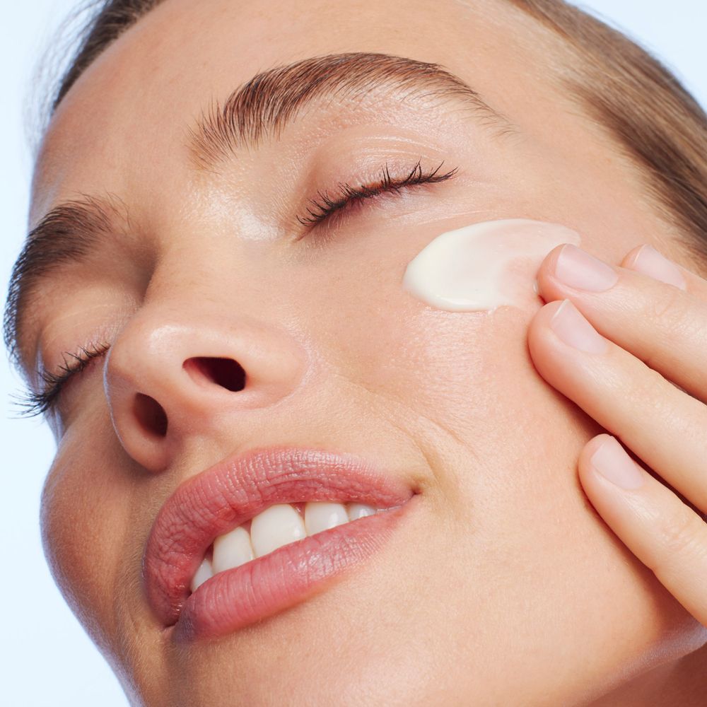 Gros plan d'une femme appliquant de la crème sur son visage. Yeux fermés, souriante.