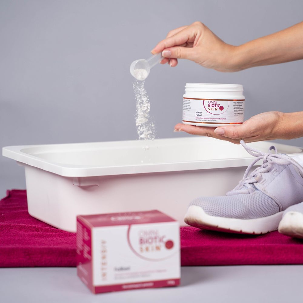Personne tenant une cuillère doseuse au-dessus d'un bain de pieds. Pot et boîte OMNi-BiOTiC SKiN® Intensiv-Fußbad.