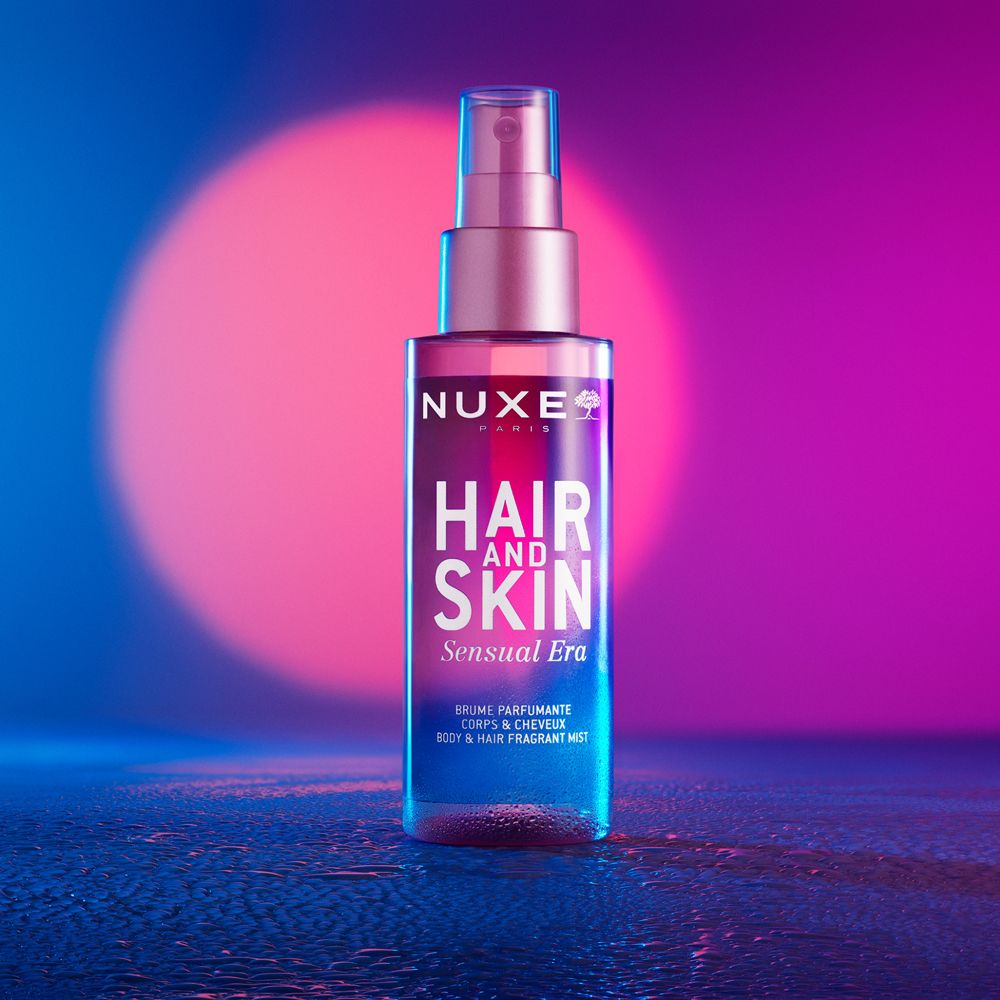 NUXE® Hair and Skin Sensual Era Duftspray. Rosa-farbene Flasche mit Sprühkopf. Blauer und pinker Hintergrund.