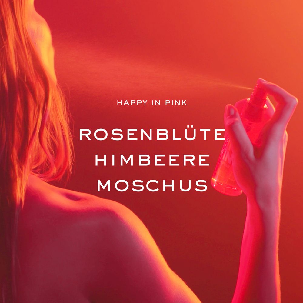 Person sprüht. Aufschrift: Happy in Pink, Rosenblüte, Himbeere, Moschus. Roter Hintergrund.