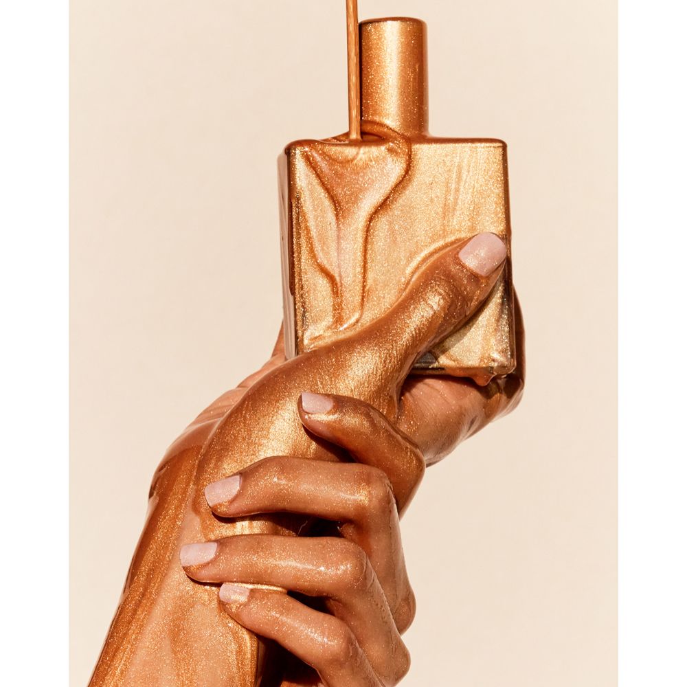 Mains tenant un flacon doré, recouvert de liquide doré. Ongles vernis.