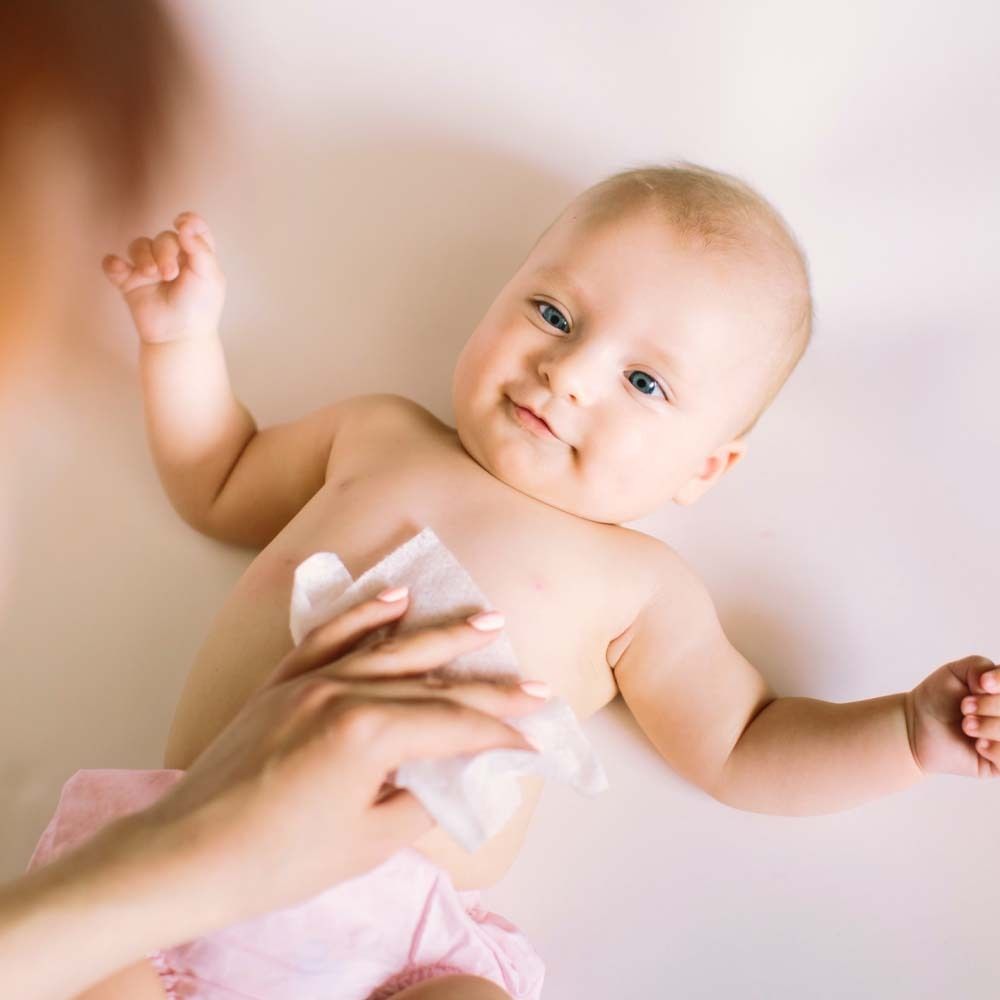 Bébé allongé sur le dos, une main nettoie le haut du corps avec une lingette blanche. Couche rose visible.