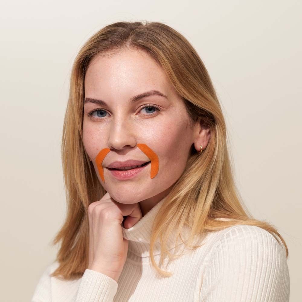 Eine Frau mit einem weißen Rollkragenpullover. Auf der Haut sind zwei orangefarbene Mund-Patches angebracht.
