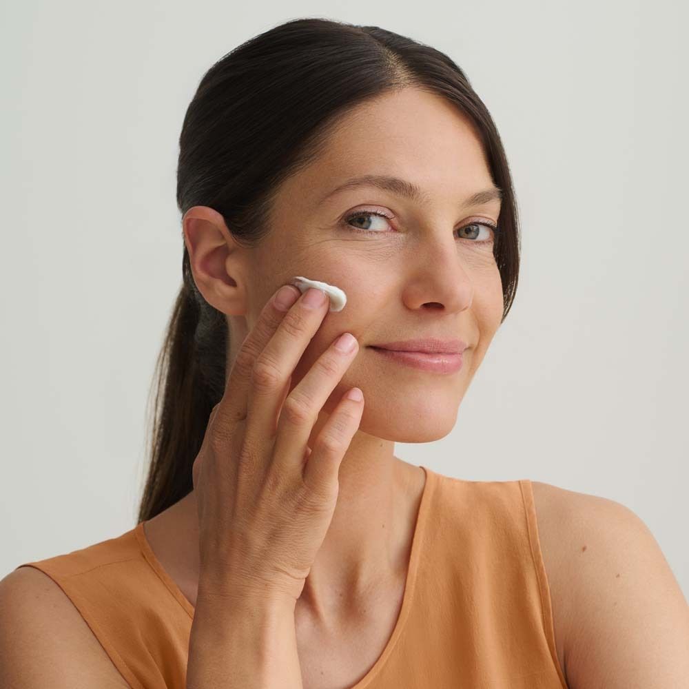 Femme appliquant de la crème sur son visage. Elle sourit. Soin de la peau. Peau claire. Haut orange. Fond clair.