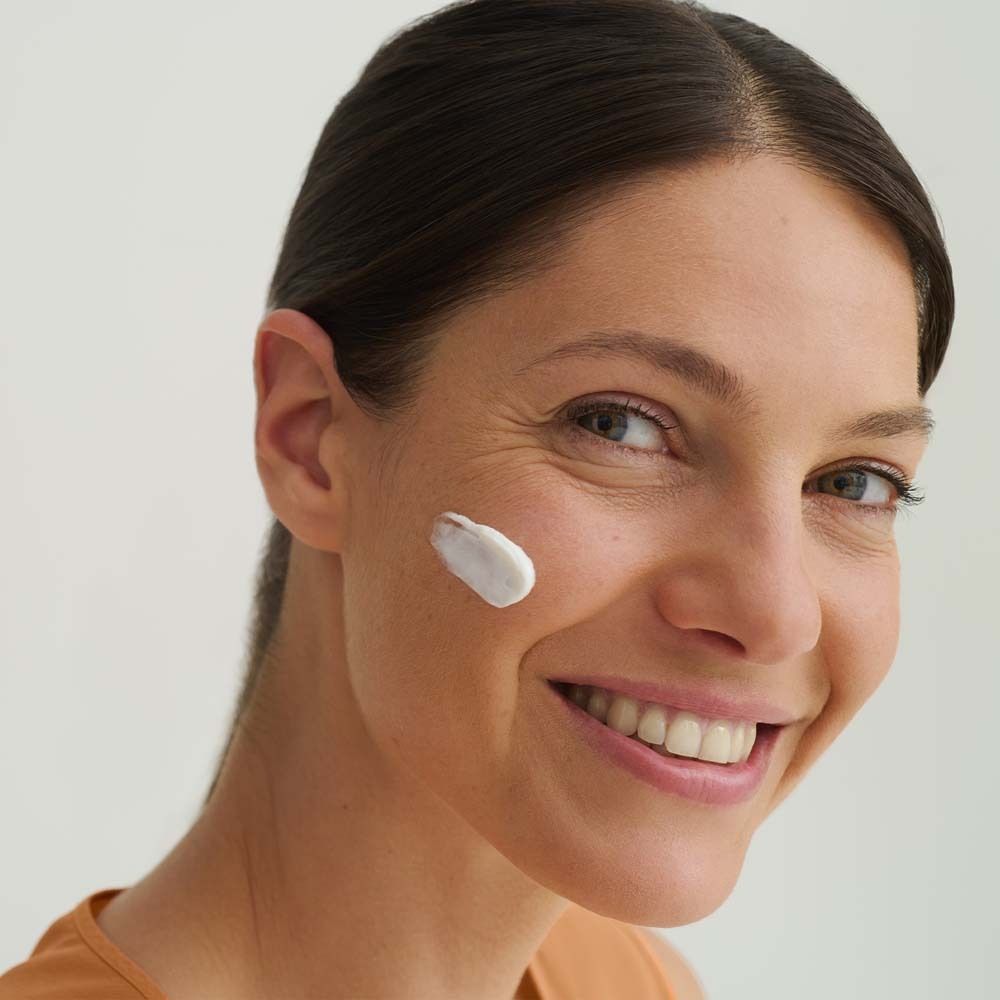 Femme avec de la crème sur la joue. Elle sourit. Soin de la peau. Peau claire. Haut orange. Fond clair.