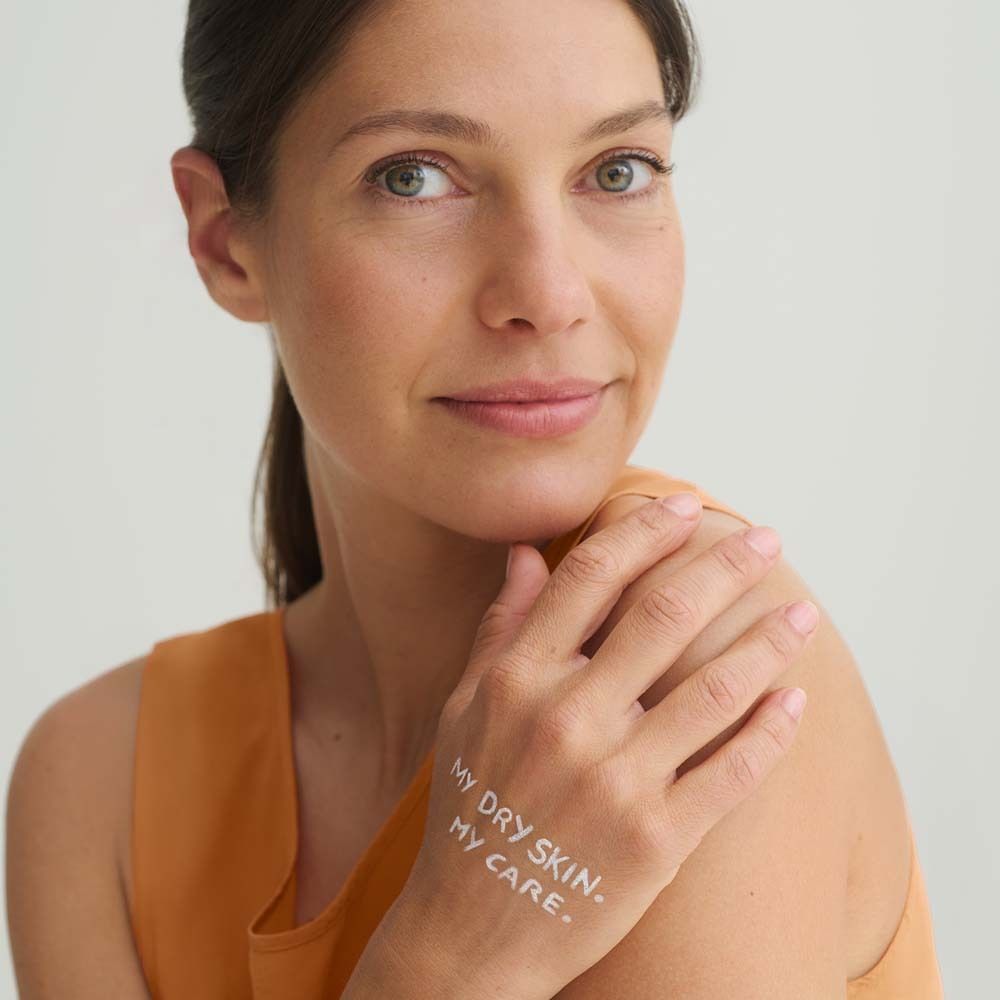 Femme avec texte sur la main : MY DRY SKIN. MY CARE. Elle porte un haut orange. Fond clair.