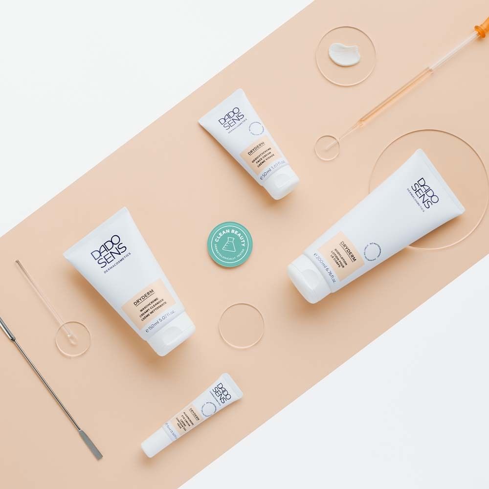 Divers produits et accessoires DADOSENS DRYDERM sur fond beige incliné. Flacons, pots, pipette, spatule.