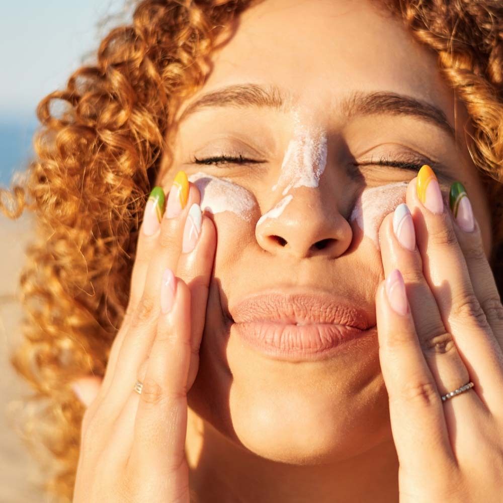Frau trägt Sonnencreme auf ihr Gesicht auf. Helle Haut, lockiges Haar, bunte Fingernägel. Nahaufnahme.