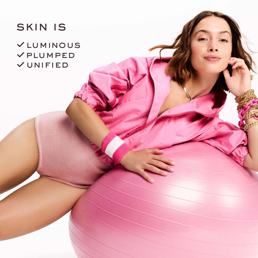 Femme allongée sur un ballon de gymnastique rose. Elle porte des vêtements de sport roses. Texte: SKIN IS, Luminous, Plumped, Unified.