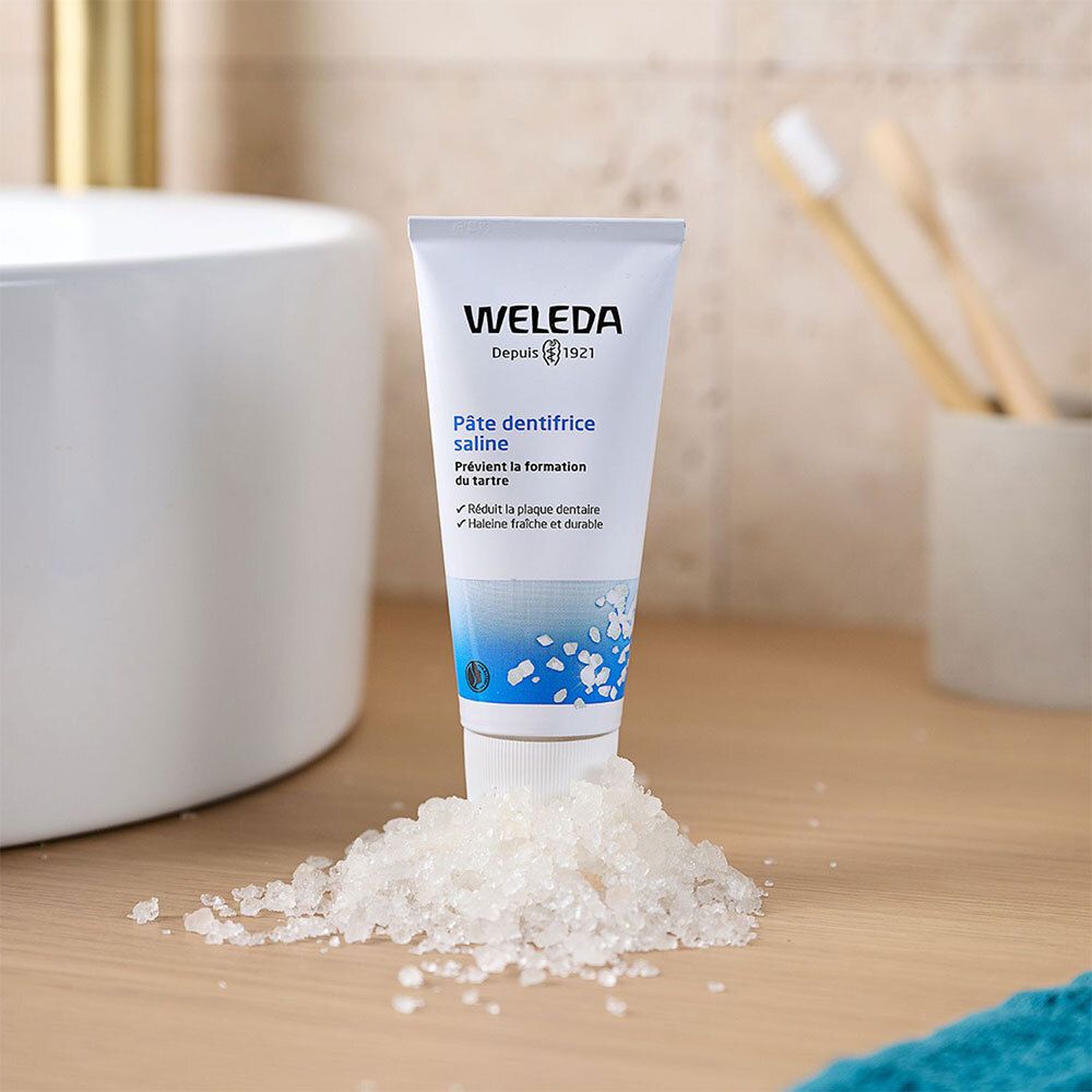 Weiße Tube mit blauer Schrift. WELEDA, Pâte dentifrice saline. Auf Tisch mit Salz. Badezimmerhintergrund.
