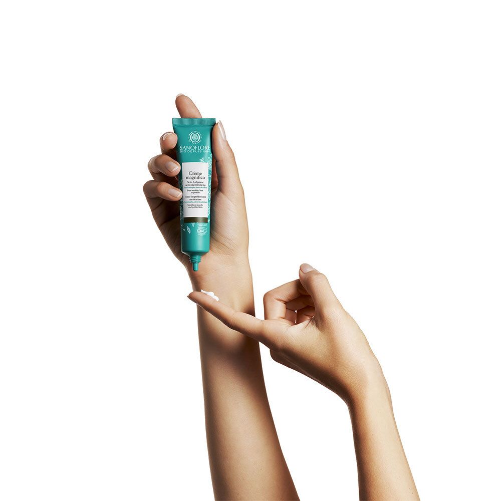 Hände halten eine türkisfarbene Tube mit Creme. Aufschrift: Crème magnifica. Creme auf Finger.