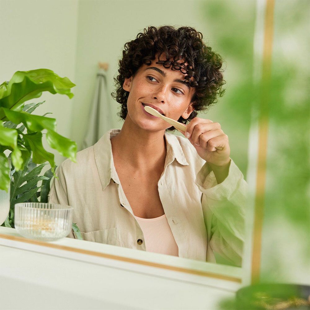 Femme se brosse les dents devant un miroir. Une plante en arrière-plan. Elle tient une brosse à dents en bois.