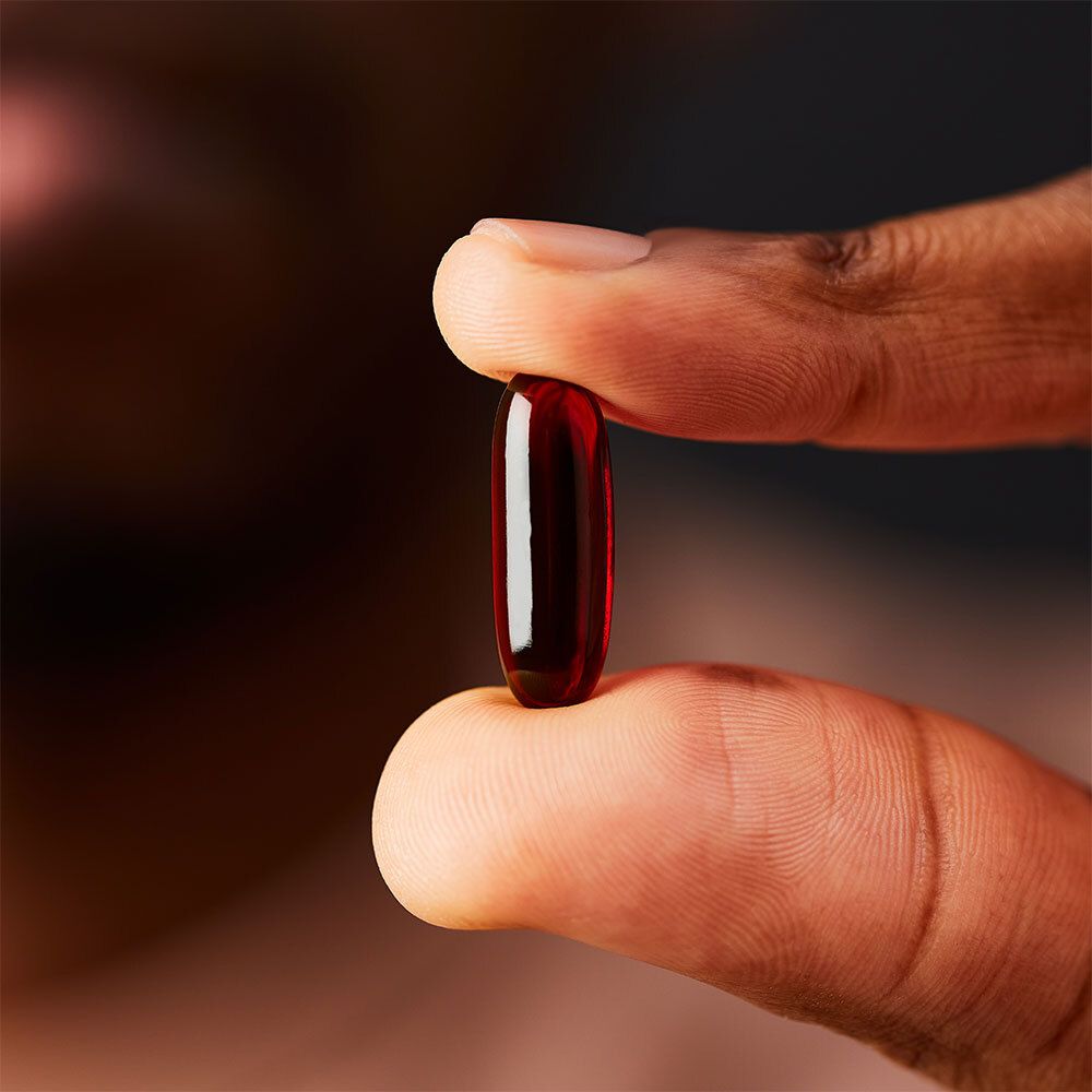 Une gélule rouge tenue entre deux doigts. Gélule ovale et transparente.