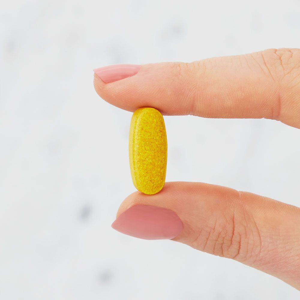 Gelbe, ovale Tablette zwischen Fingern gehalten. Nahaufnahme, heller Hintergrund.