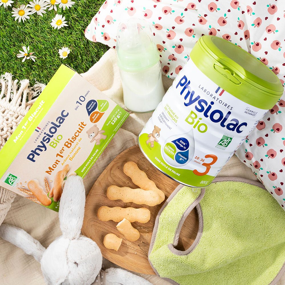 Szene mit Physiolac Bio Produkten. Mon 1er Biscuit Banane, Milchpulver, Flasche, Lätzchen und Spielzeug auf Gras.