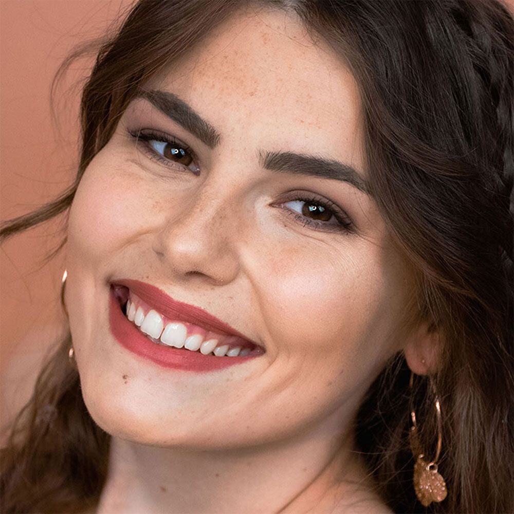 Porträt einer Frau mit geformten Augenbrauen. Sie trägt Lippenstift und Ohrringe. Haut mit Sommersprossen.