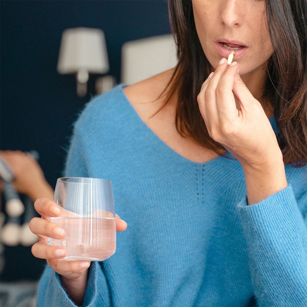 Frau nimmt eine Kapsel ein. Sie hält ein Glas Wasser. Blaues Oberteil.