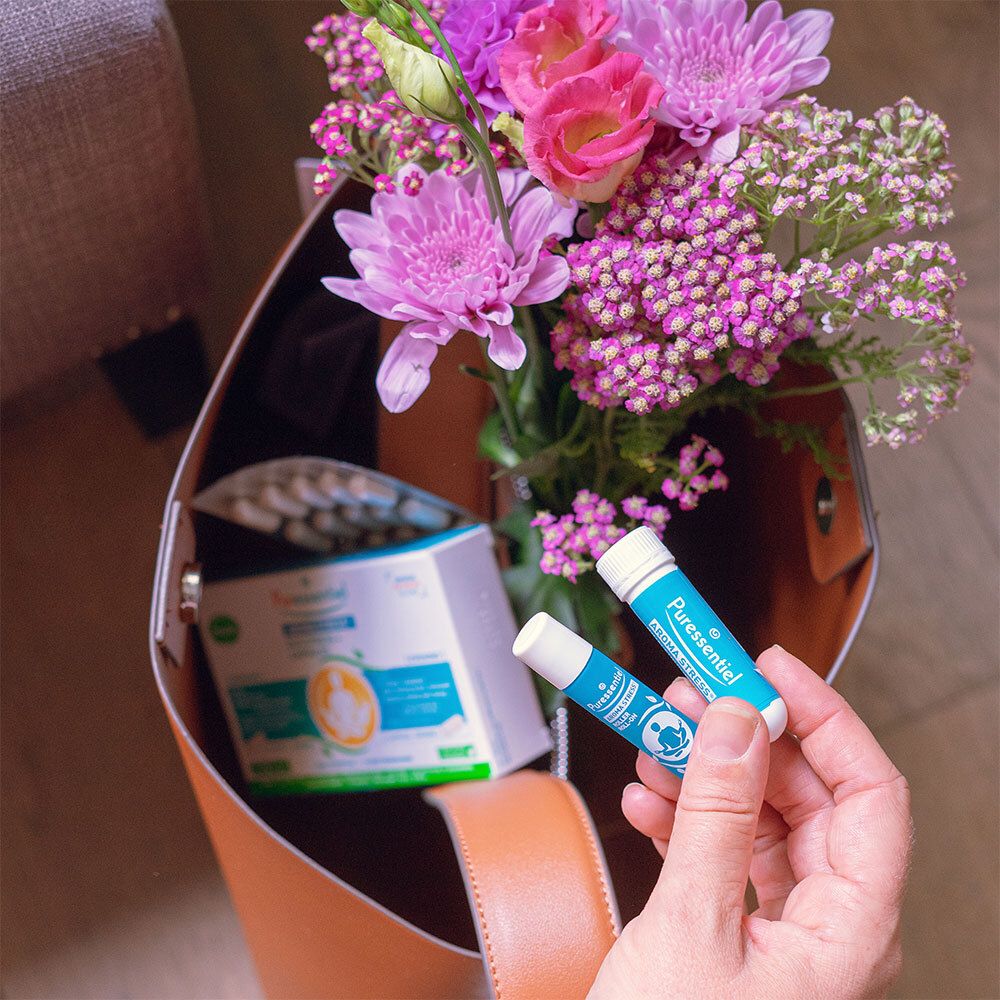 Hand hält zwei blaue Stifte und eine Packung Puressentiel AROMA STRESS®. Blumen und Tasche.