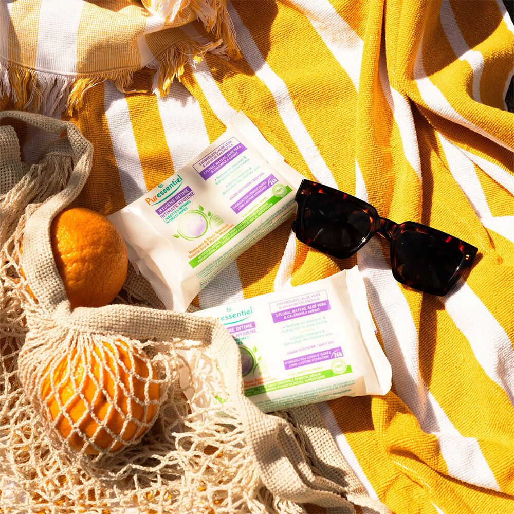 Zwei Packungen Intim-Reinigungstücher auf einem gelb-weiß gestreiften Handtuch. Text: HYGIÈNE INTIME. Sonnenbrille und Netzbeutel mit Orange.