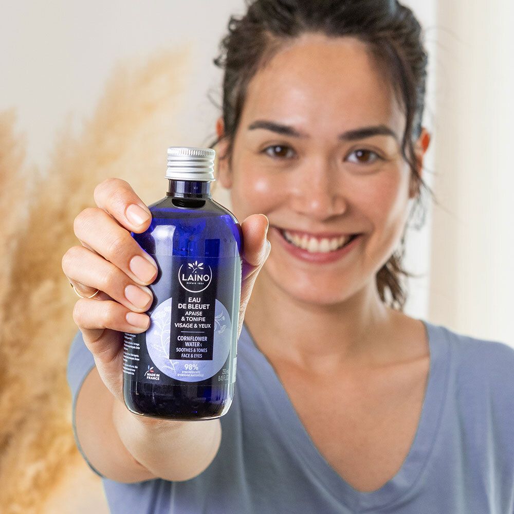 Femme tenant un flacon LAINO Eau de Bleuet. Flacon bleu avec bouchon argenté. Inscription: Eau de Bleuet, Cornflower Water.