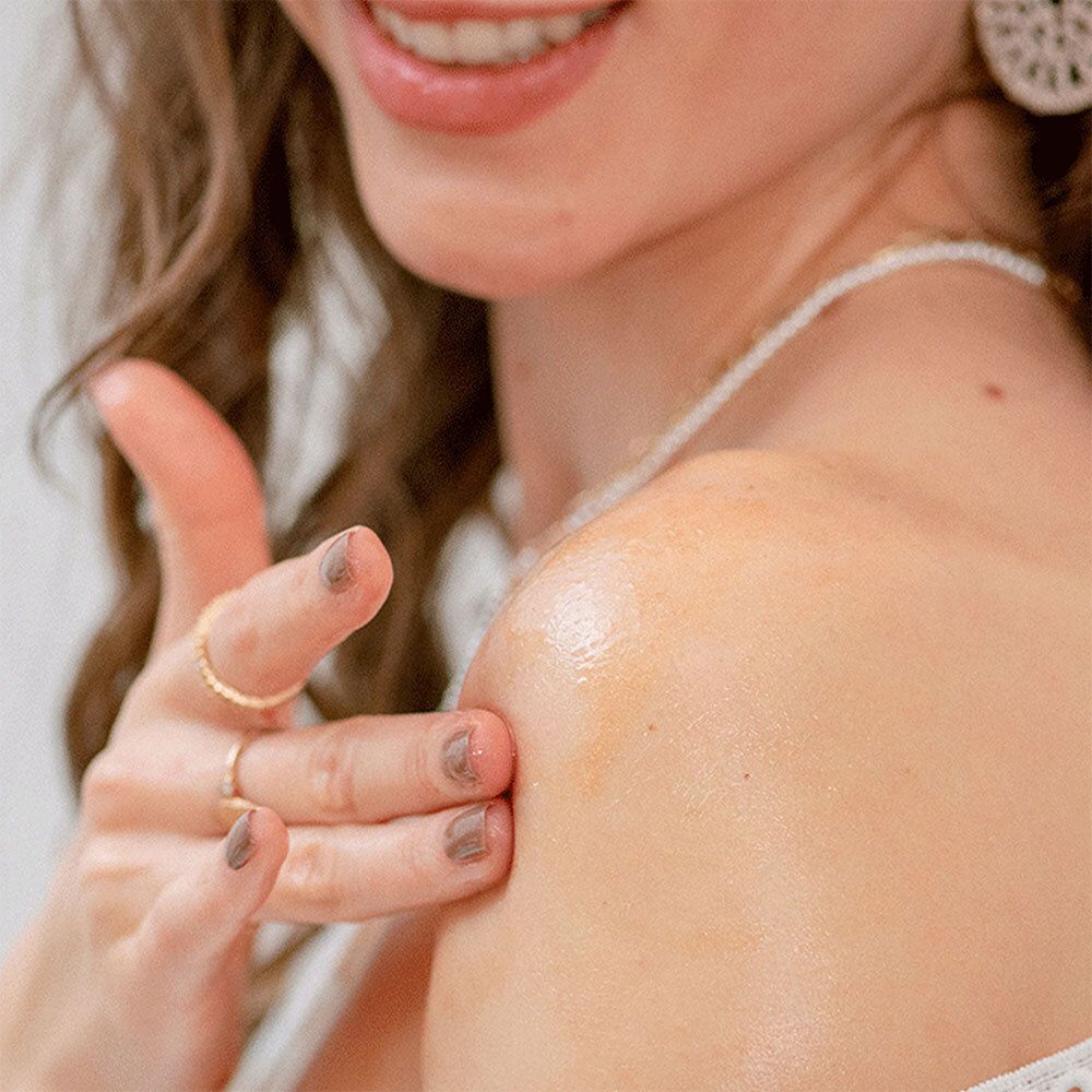 Person trägt Schimmeröl auf die Schulter auf. Goldfarbenes Öl. Helle Haut. Ringe an der Hand.