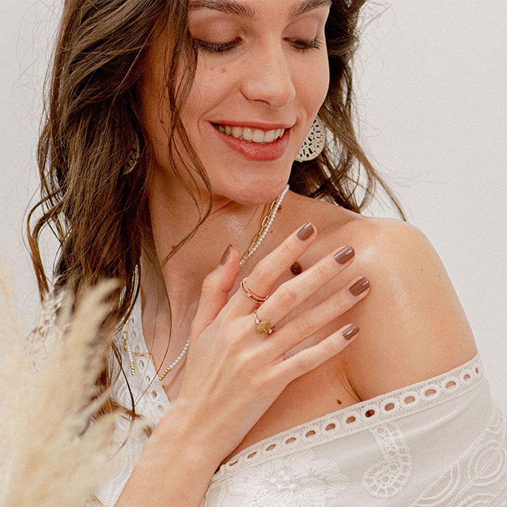 Frau mit braunen Haaren, trägt Schmuck. Sie berührt ihre Schulter. Helle Kleidung.