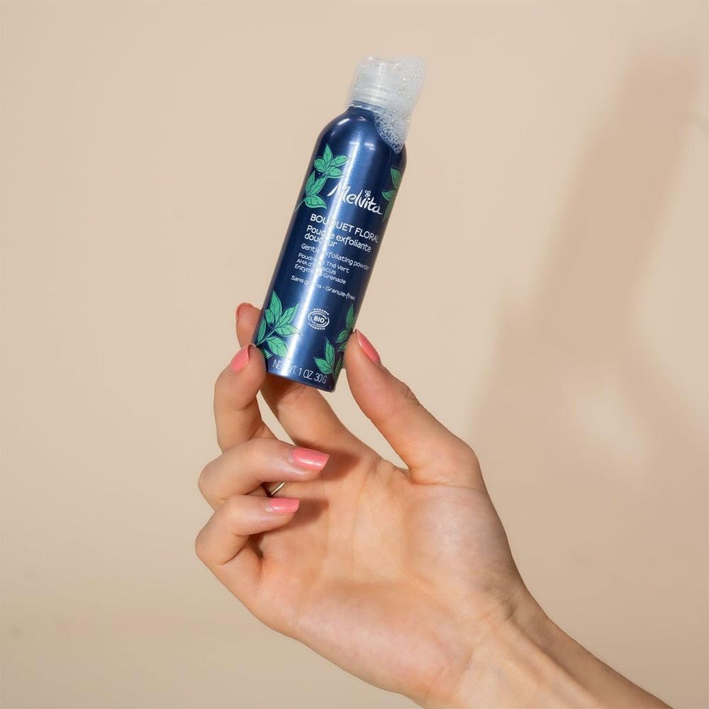 Hand hält blaue Flasche mit weißem Deckel. Aufschrift: Melvita, Bouquet Floral, Poudre exfoliante douceur. Bio-Zertifizierung.