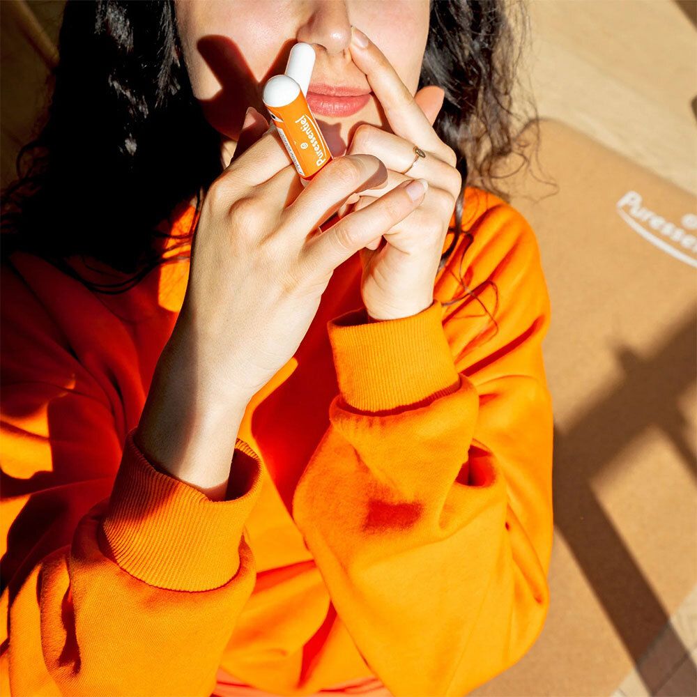 Eine Person hält einen weißen und orangefarbenen Inhalator vor ihrer Nase. Sie trägt einen orangefarbenen Pullover.
