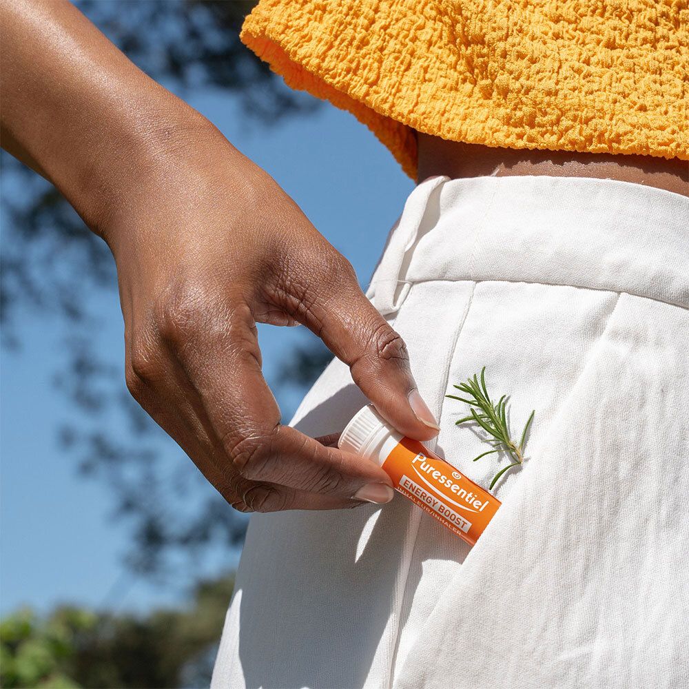 Eine Hand hält einen weißen und orangefarbenen Inhalator in einer Hosentasche. Ein Zweig Rosmarin liegt daneben.