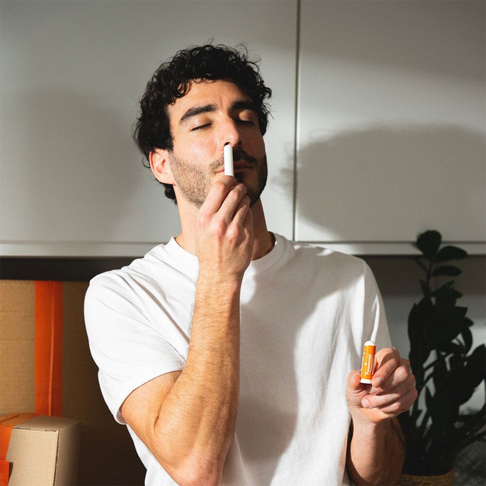 Ein Mann hält einen weißen und orangefarbenen Inhalator vor seiner Nase. Er trägt ein weißes T-Shirt.