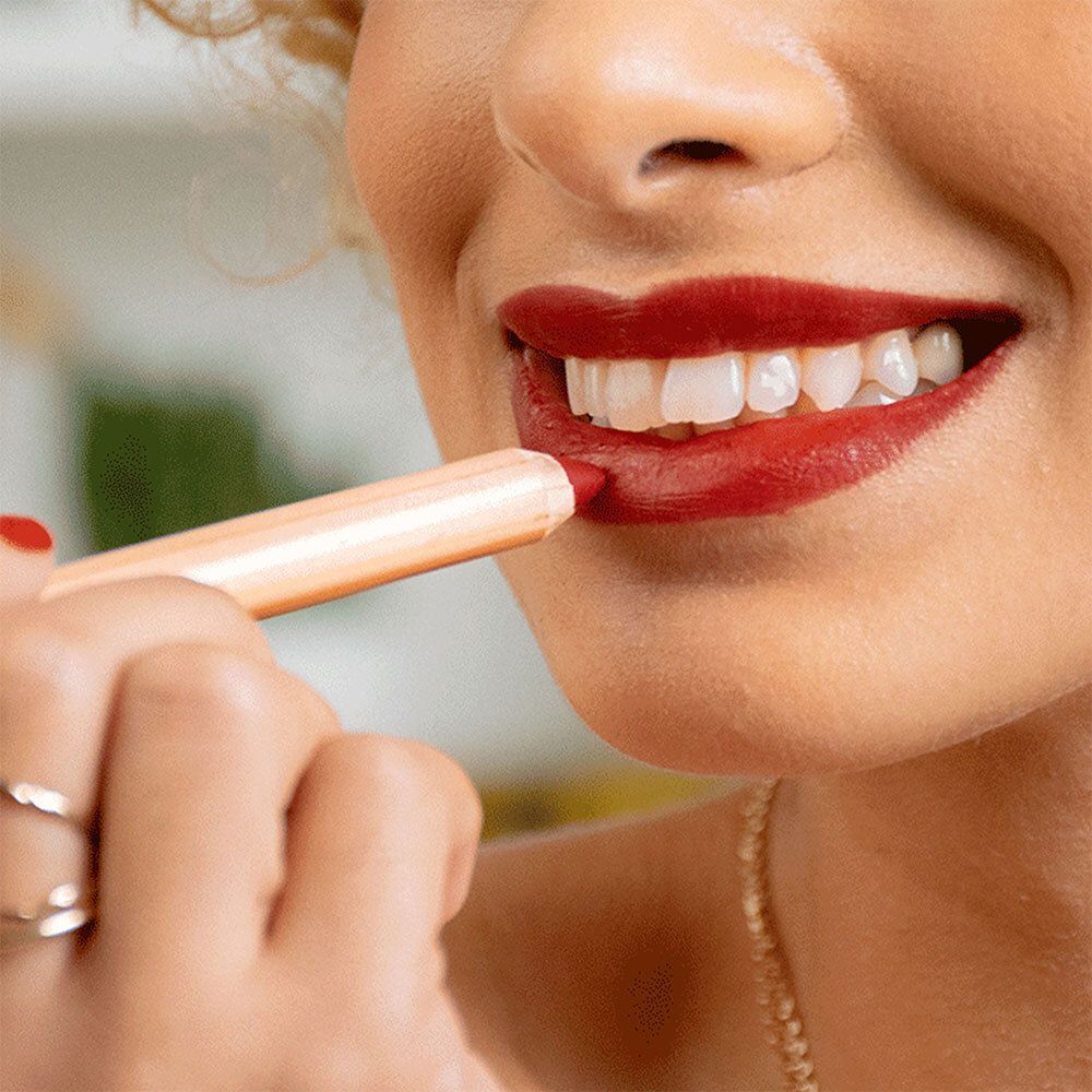 Person trägt roten Lippenstift auf. Der Lippenstift ist in einem hellen Holzton gehalten.