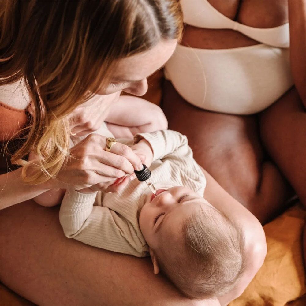 Eine Person tropft Flüssigkeit aus einer Pipette in den Mund eines Babys. Das Baby liegt auf dem Schoß einer anderen Person.