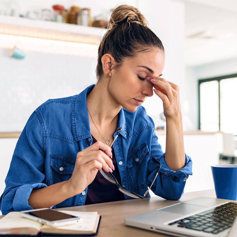 Femme aux yeux fermés, main sur la tempe, assise à un bureau.