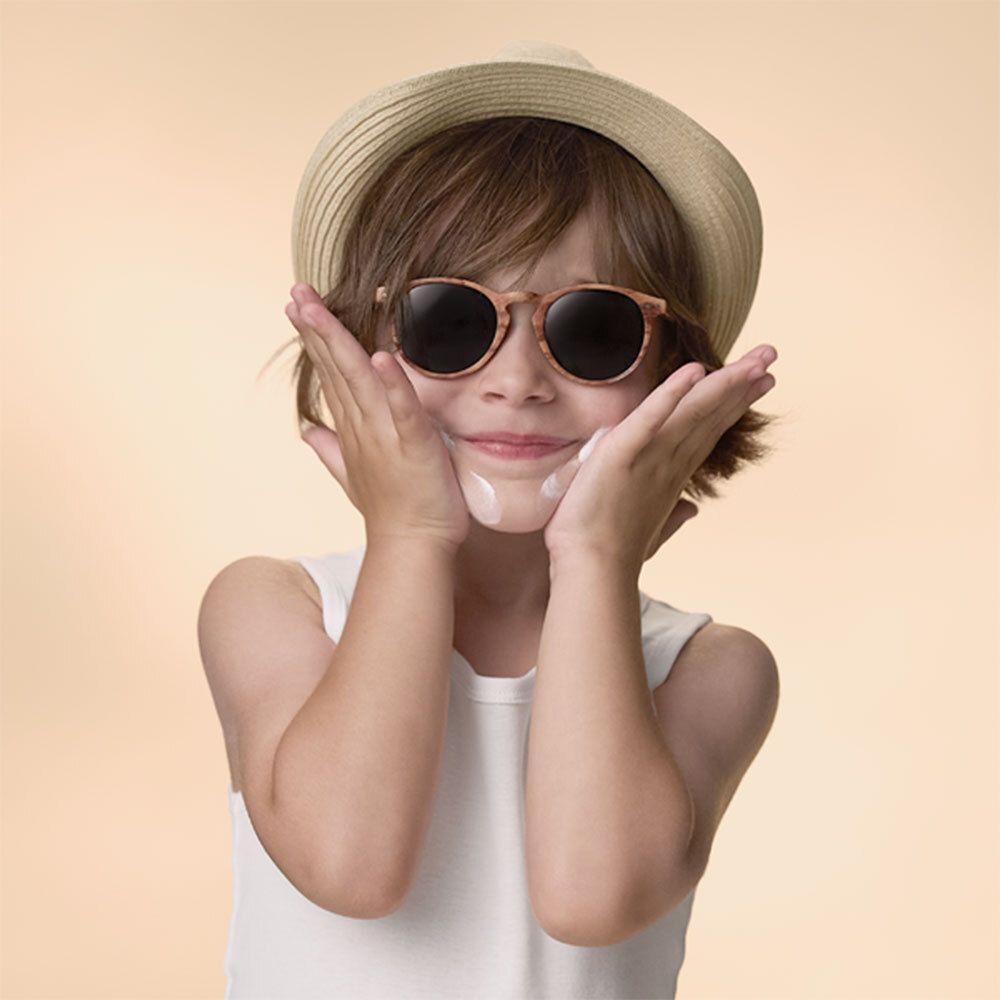 Enfant avec lunettes de soleil et chapeau. Crème sur les joues. Mains sur le visage.