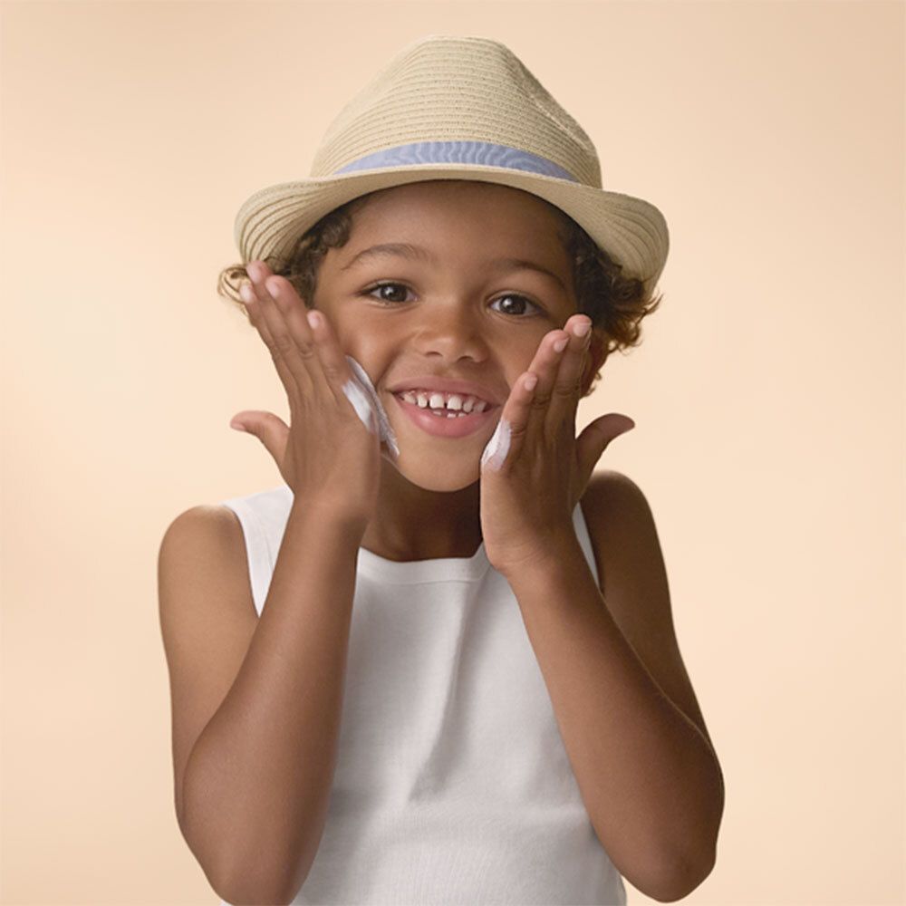 Enfant appliquant crème solaire sur le visage. Chapeau et débardeur. Crème sur les mains.