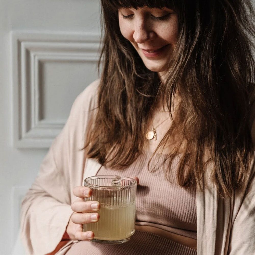 Femme enceinte tenant un verre de boisson jaune. Elle sourit. Arrière-plan : pièce blanche.