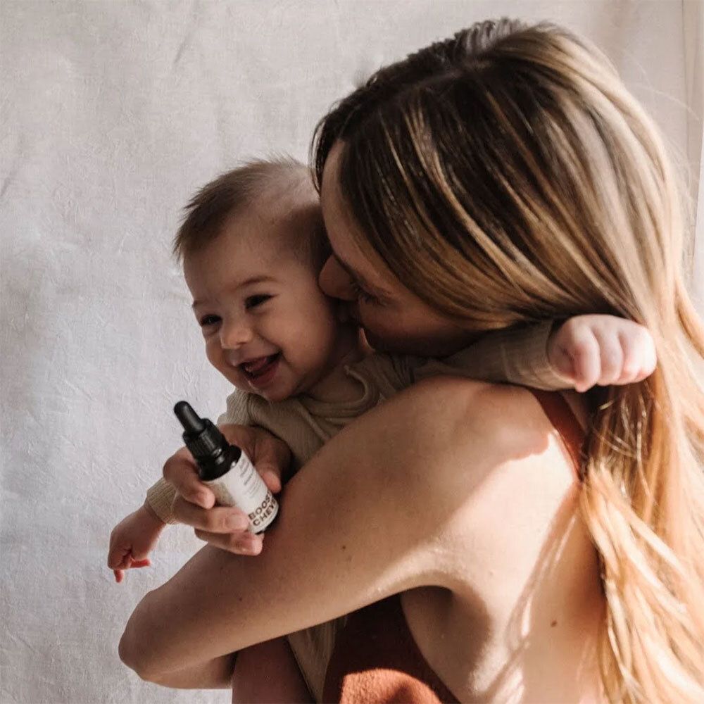 Frau hält Baby im Arm. Baby lächelt. Frau hält eine Flasche mit Tropfer.