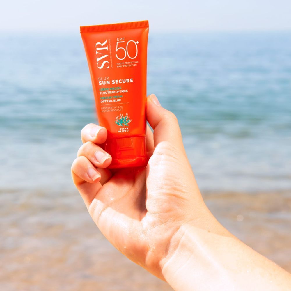Main tenant un tube de SVR Sun Secure Blur LSF50+. Tube orange avec texte blanc. Mer en arrière-plan.
