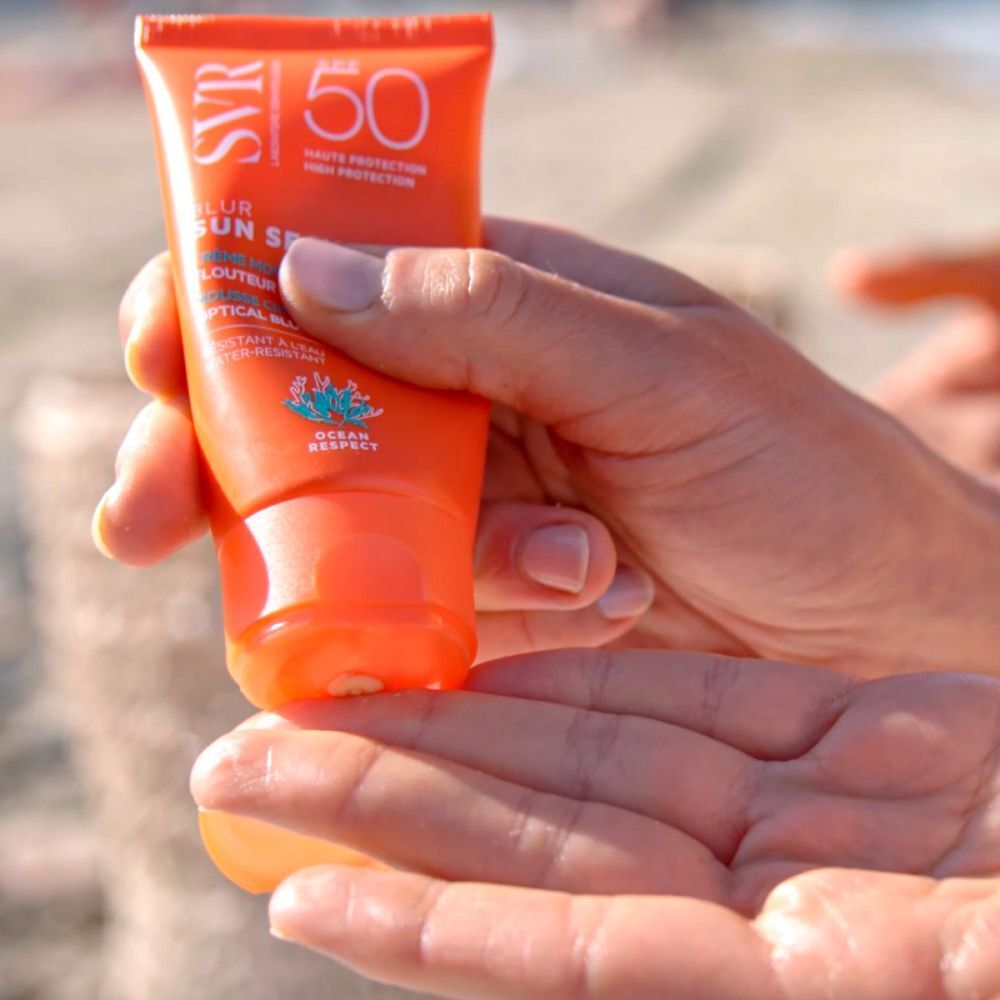 Main tenant un tube de SVR Sun Secure Blur LSF50+. Tube orange avec texte blanc. Mer en arrière-plan.