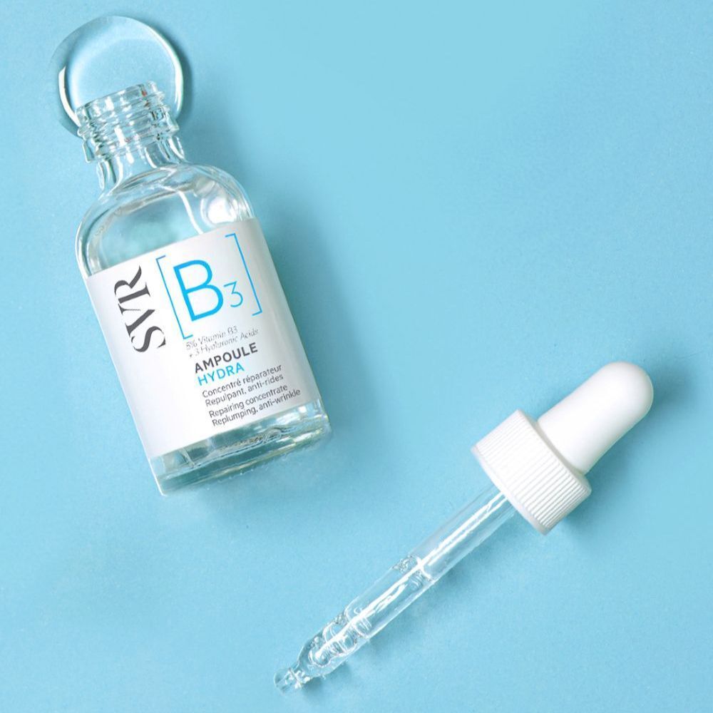 Une personne tient un flacon SVR [B3] Ampoule Hydra. Carrelage bleu en arrière-plan.