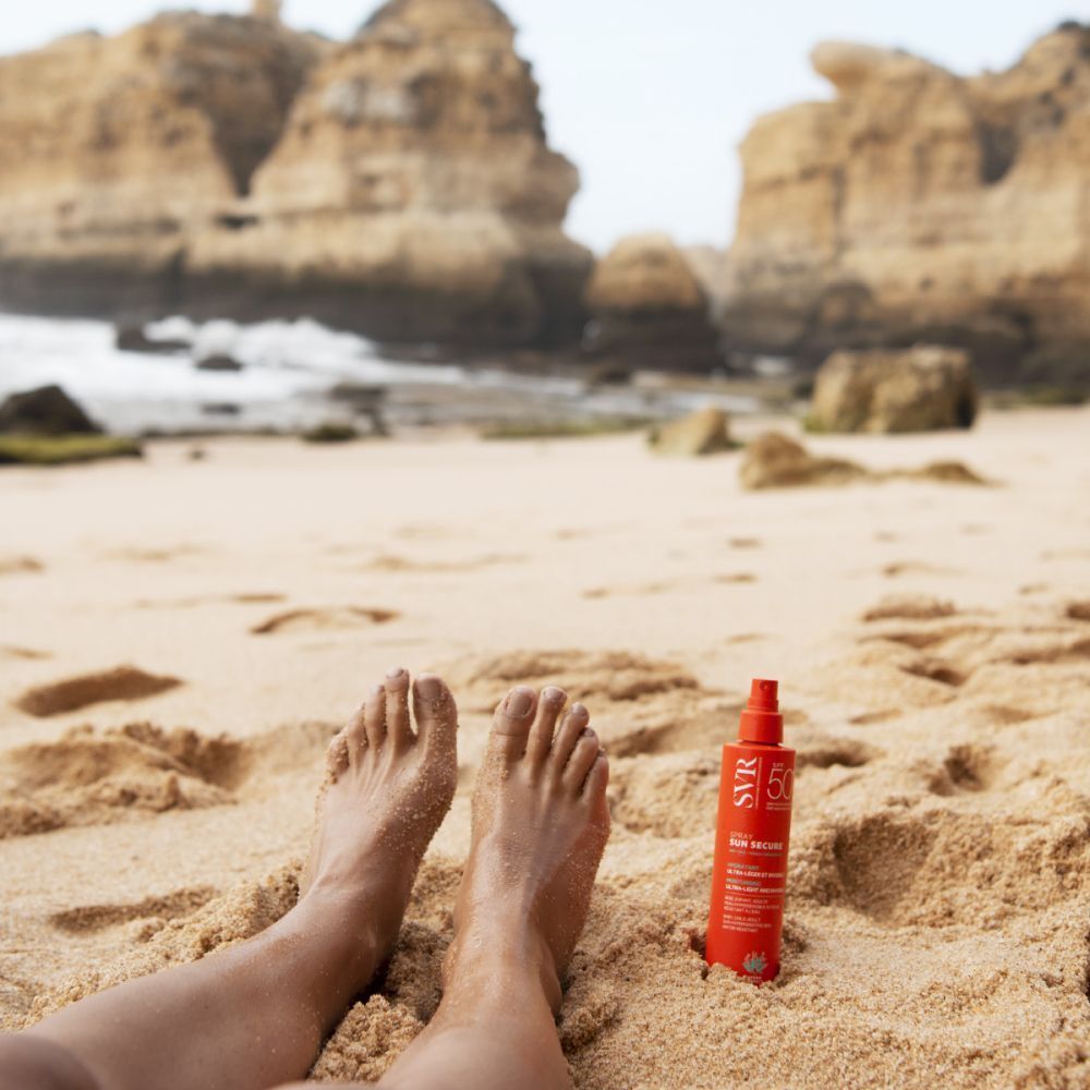 Un flacon orange est posé sur la plage à côté de pieds. Inscription : SVR, SUN SECURE, SPF 50+.