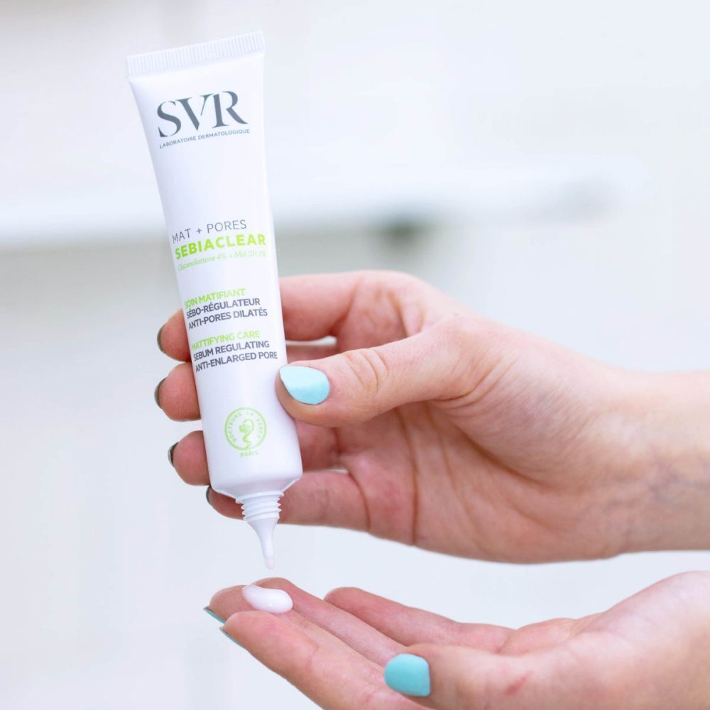 Tube de SVR Sebiaclear Matte + Poren pressé dans une main. Crème blanche pressée dans l'autre main. Peau claire, ongles bleus.