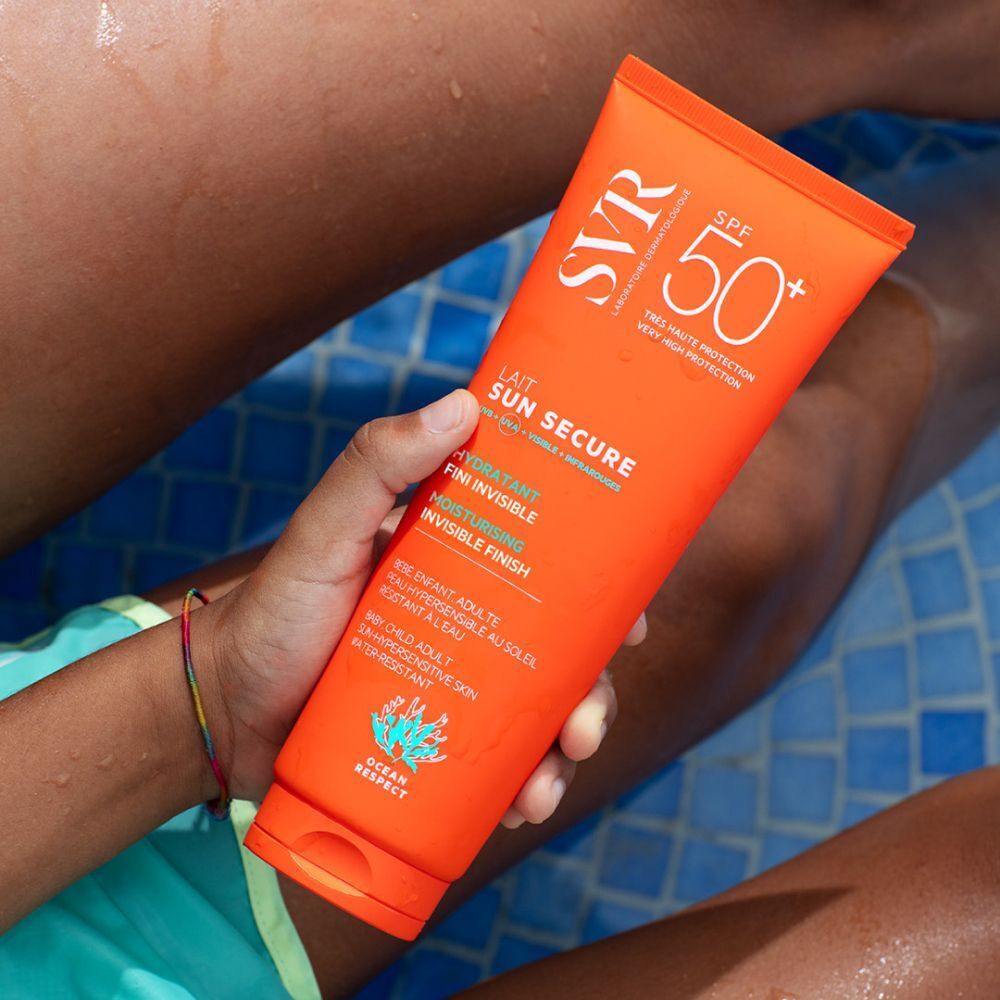 Personne tenant un tube de SVR SUN SECRUE Lait LSF 50+. Tube orange avec texte blanc. Texte: Lait Sun Secure, Fini Invisible, Moisturising.